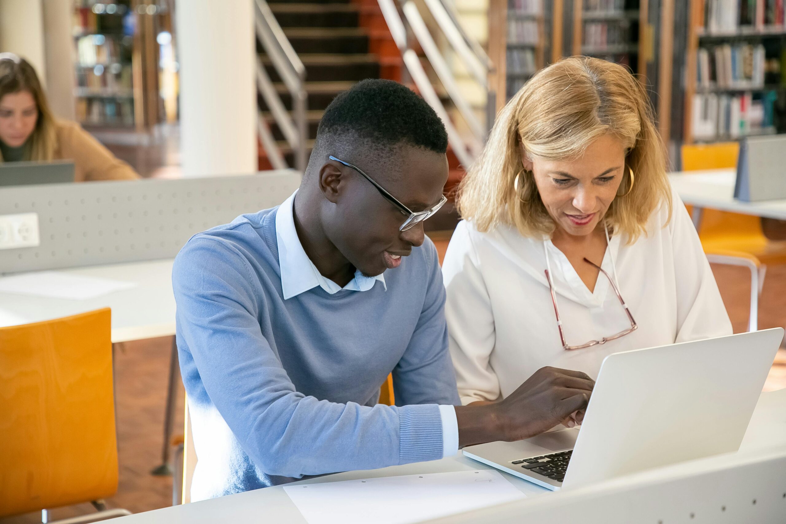 Lehrerin Hilft Ethnischen Männlichen Studenten, Die Am Laptop In Der Bibliothek Arbeiten